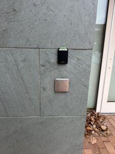 The automatic door opener is a silvery/grey color with no lettering on it located below the Pcard reader. It is located to the right of the doors on the wall made of grey slate slabs. 