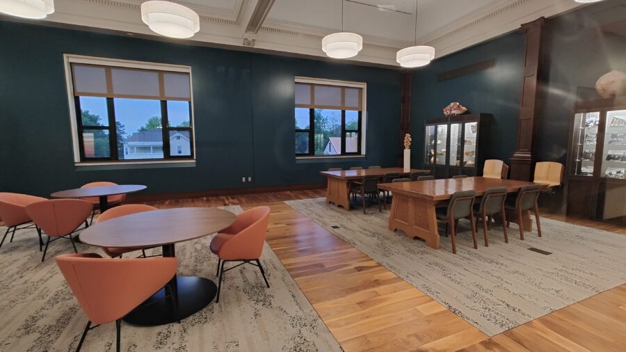 View of the northern half of the Faculty/Staff Lounge, showing 2 exterior windows, seating area with desk-height round tables and study-desks.